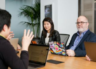 What Small Business Owners Need To Know About Workers Compensation Insurance in Pennsylvania boardroom meeting where woman in black leather jacket and man in blue checkered shirt, suit jacket and glasses, pay attention to person talking with their hands wearing black long sleeve shirt
