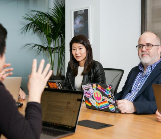 boardroom meeting where woman in black leather jacket and man in blue checkered shirt, suit jacket and glasses, pay attention to person talking with their hands wearing black long sleeve shirt