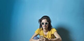 Photo of Woman in Yellow T-shirt, Blue Jeans, and White Sunglasses Sitting on Wooden Floor In Front of Blue Wall