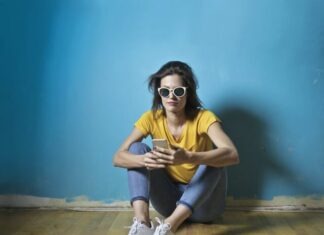 Photo of Woman in Yellow T-shirt, Blue Jeans, and White Sunglasses Sitting on Wooden Floor In Front of Blue Wall