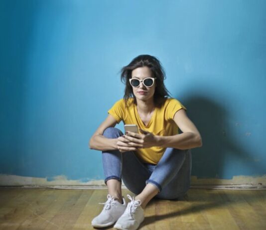 Photo of Woman in Yellow T-shirt, Blue Jeans, and White Sunglasses Sitting on Wooden Floor In Front of Blue Wall