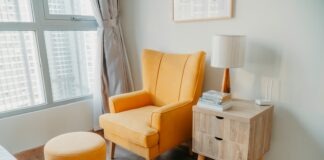 yellow armchair and stool beside wooden nightstand by the wall near glass window and bed