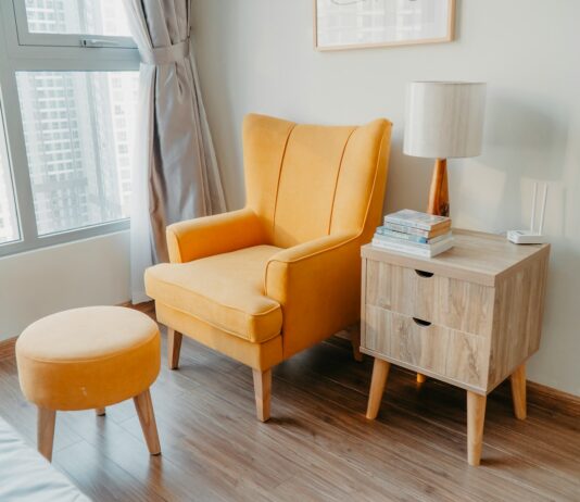 yellow armchair and stool beside wooden nightstand by the wall near glass window and bed