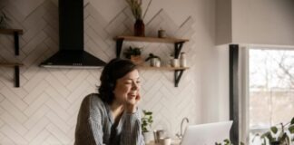 Young cheerful female smiling and talking via laptop while sitting at wooden table in cozy kitchen