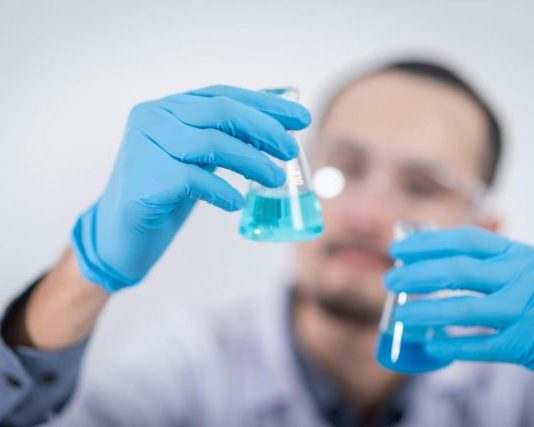 Scientist in a laboratory conducting an experiment, holding blue liquid in flasks with protective gloves.