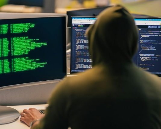 Hacker in hoodie working on multiple computer screens