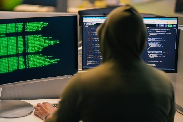 Photo by Julio Lopez Hacker in hoodie working on multiple computer screens